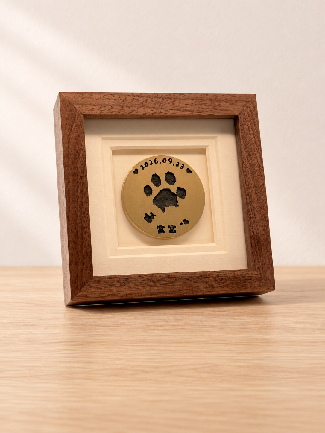 A square wooden frame with a brass paw-print disc on a pale oak table against a cream wall.