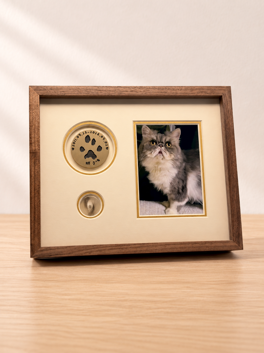 A wooden frame with a paw-print disc, cat photo and fur window on a pale oak table against a cream wall.